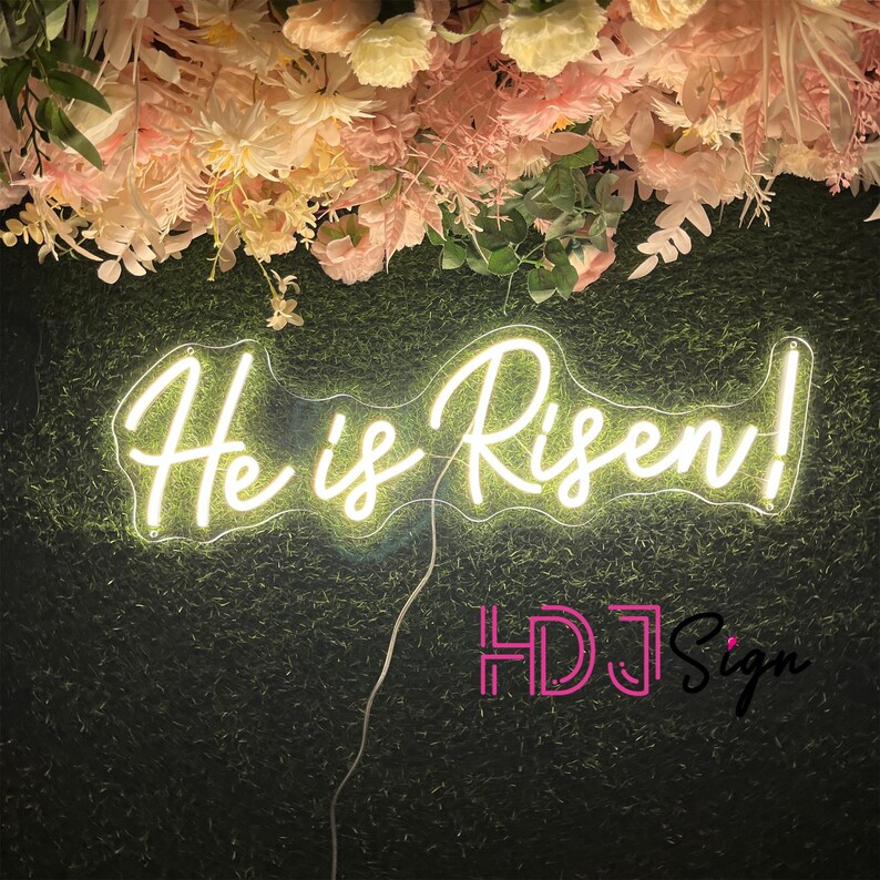 May include: White neon sign with the words "He is Risen!" on a green background with pink flowers above the sign. The sign is lit up and the words are in a cursive font.