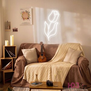 May include: A white neon sign shaped like a tulip flower, hanging on a wall above a brown couch with a white throw blanket and a ukulele. The couch is in a living room with a wooden shelf with candles and books.