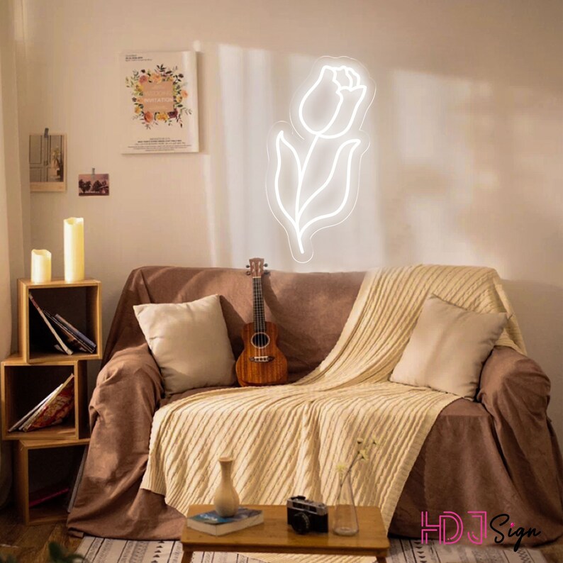 May include: A white neon sign shaped like a tulip flower, hanging on a wall above a brown couch with a white throw blanket and a ukulele. The couch is in a living room with a wooden shelf with candles and books.
