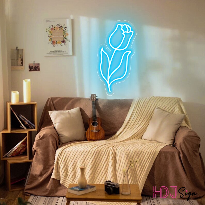 May include: A blue neon sign shaped like a tulip flower, hanging on a wall above a brown couch with a throw blanket and pillows. A ukulele is sitting on the couch.