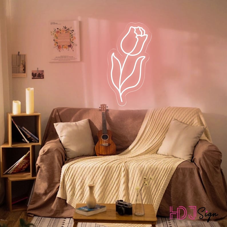 May include: A white neon sign shaped like a tulip flower with a pink glow. The sign is hanging on a pink wall above a brown couch with a white throw blanket and a ukulele.
