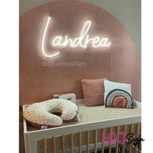 Op de afbeelding: Een wit neon-bord met de naam "Andrea" in een cursief schrift, gemonteerd op een roze muur. Het bord is verlicht en geeft een zacht gloed. Het bord maakt deel uit van een kinderkamerdecorset, met een wit wiegje en een roze en wit gepatroneerd kussen.