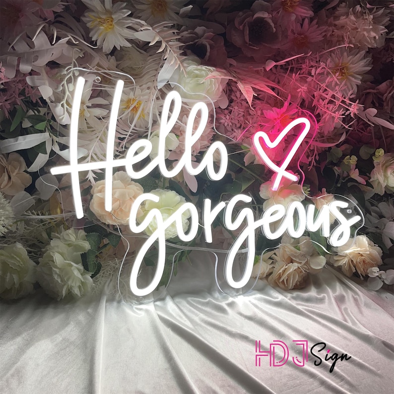 May include: White neon sign with the text "Hello gorgeous" and a pink heart. The sign is on a white background with flowers.