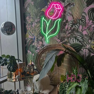 May include: A pink neon light shaped like a rose with green leaves against a pink and green floral wallpaper. A wicker chair with a white cushion sits in front of the light.