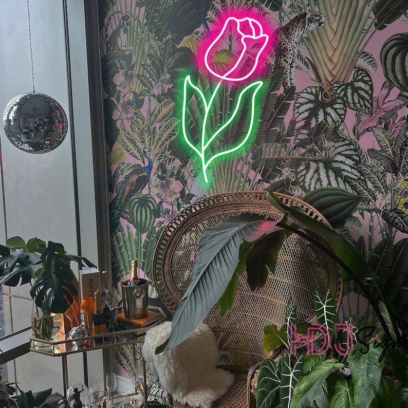 May include: A pink neon light shaped like a rose with green leaves against a pink and green floral wallpaper. A wicker chair with a white cushion sits in front of the light.