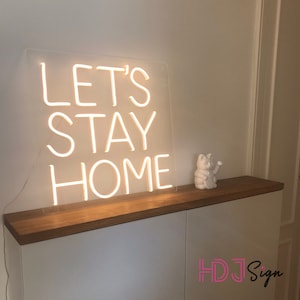 May include: White neon sign with the words "Let's Stay Home" on a clear acrylic background. The sign is sitting on a wooden shelf with a white ceramic figurine of a cat.