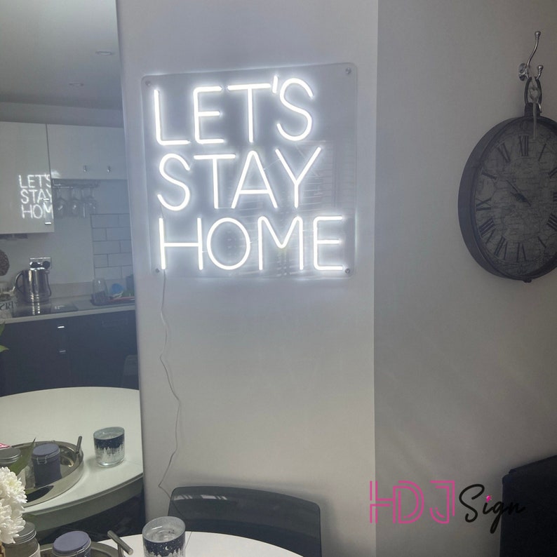 May include: White neon sign with the words "Let's Stay Home" on a white wall. The sign is lit up and the letters are outlined in white.
