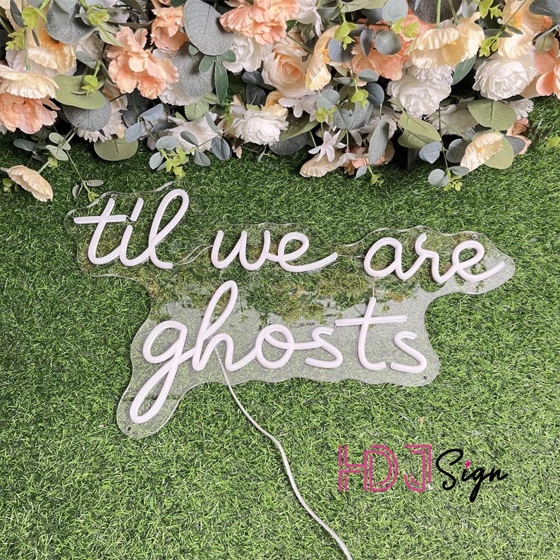 May include: A clear acrylic sign with the words "til we are ghosts" in white script. The sign is on a bed of green moss and grass, with a floral arrangement of peach and white flowers in the background.