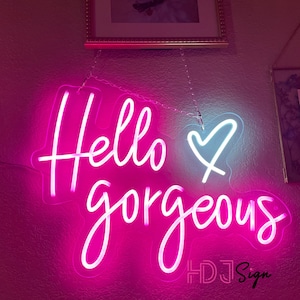 May include: Pink neon sign with the text "Hello gorgeous" and a white heart. The sign is hanging from a chain and has the text "HDJ Sign" in the bottom right corner.