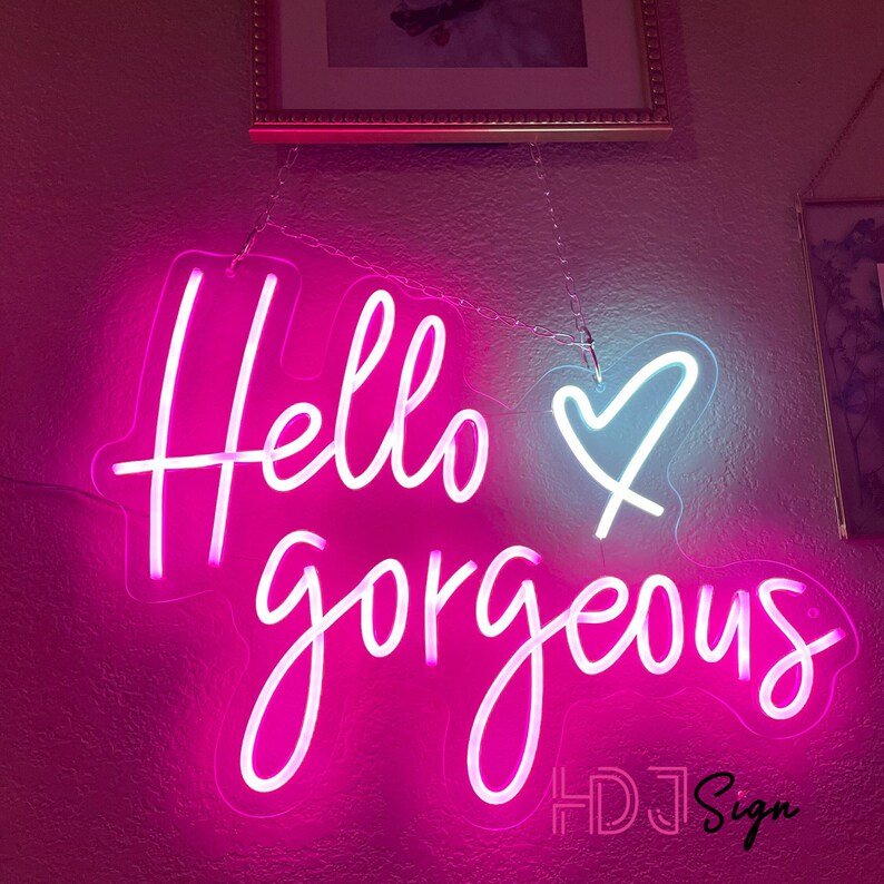 May include: Pink neon sign with the text "Hello gorgeous" and a white heart. The sign is hanging from a chain and has the text "HDJ Sign" in the bottom right corner.