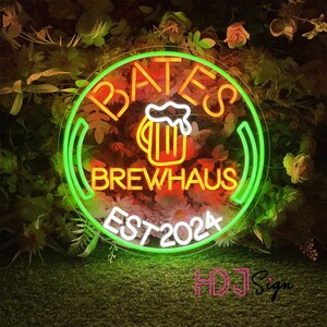 May include: Neon sign with a green circle and white text that reads "Bates Brewhaus Est 2024". Inside the circle is a yellow beer mug with orange liquid and white foam. The sign is hanging in front of a green leafy background.