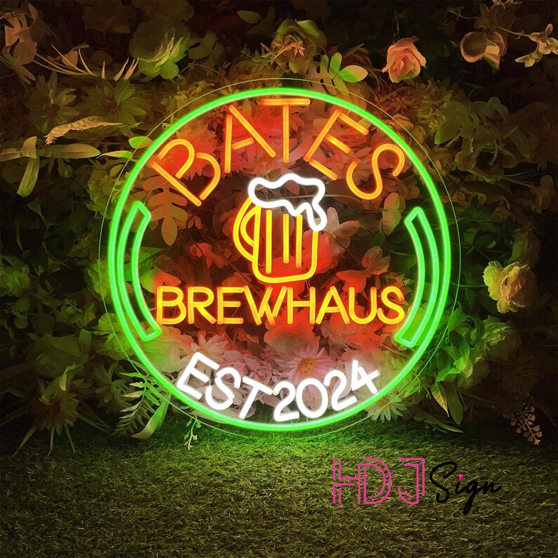 May include: Neon sign with a green circle and white text that reads "Bates Brewhaus Est 2024". Inside the circle is a yellow beer mug with orange liquid and white foam. The sign is hanging in front of a green leafy background.