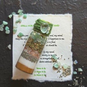 Op de afbeelding: Een klein glazen flesje gevuld met groene en roze kristallen, kruiden en glitter. Het flesje is vastgebonden met een groene touwtje en heeft een groen en wit etiket met de tekst "SPELL FOR AND" en een gedicht over geluk en kracht.