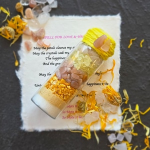 May include: A glass bottle with a yellow string tied around the top, filled with small pink, yellow, and white crystals, and a piece of paper with the text "SPELL FOR LOVE & HAPPINESS" written on it. The bottle is sitting on a white piece of paper with the text "May the petals cleanse my soul, May the crystals seek my happiness, The happiness, And the great, May the, Unt, Mote it, So Mote it, So Mote it be, happiness to be" written on it.