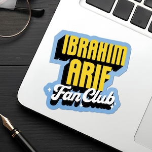 Puede incluir: Una pegatina en un portátil con el texto "IBRAHIM ARIF Fan Club" en amarillo y blanco, delineado en negro y azul. La pegatina está en un portátil blanco, con un teclado negro. También se ven unas gafas con montura dorada y un bolígrafo.