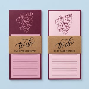 May include: Two DL 50 page notepads with lined pages. One notepad has a burgundy cover with the words "Always Shit to-do" in cursive. The other notepad has a pink cover with the same text. Both have a brown band that says "to do".