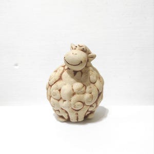 Puede incluir: Figura de oveja de cerámica beige con una cara sonriente y un cuerpo texturizado. La oveja tiene forma redonda y está sobre una superficie blanca. La figura es un objeto decorativo.