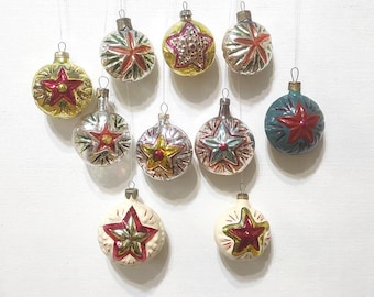 Puede incluir: Una colección de adornos navideños vintage. Los adornos son redondos, con varios colores como dorado, plateado y verde azulado. Cada adorno presenta un diseño de estrella en rojo, verde y dorado. Están suspendidos por finos hilos sobre un fondo blanco.
