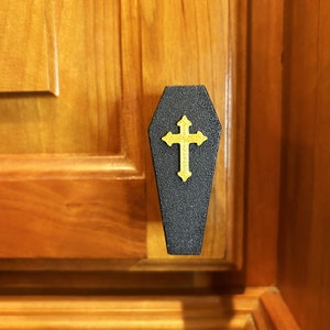May include: A black coffin-shaped cabinet knob with a gold cross on the front. The knob is mounted on a wooden cabinet door.
