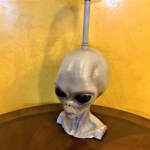 May include: A gray alien head lamp with a silver metal lampshade. The alien head is sculpted with realistic details and has large black eyes.
