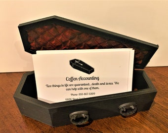 May include: A black coffin-shaped box with a red interior and a business card inside. The business card reads "Coffen Accounting" and includes the tagline "Two things in life are guaranteed...death and taxes. We can help with one of them." The card also includes a phone number and address.