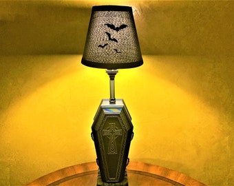 May include: A table lamp with a black lampshade featuring a bat design. The base of the lamp is shaped like a coffin with a Celtic cross.