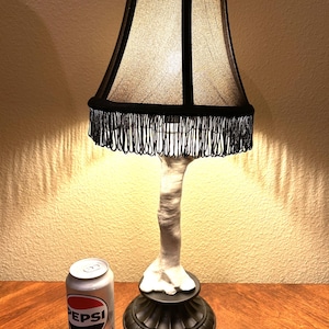 May include: A white lamp with a black lampshade and fringe. The lamp is shaped like a leg and foot. A can of Diet Pepsi is in the foreground.