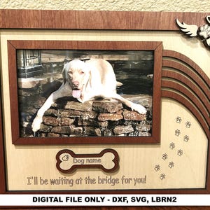 Puede incluir: Un marco conmemorativo de madera para mascotas con una foto de un perro. El marco es marrón con un interior de madera clara. Incluye una etiqueta con forma de hueso, huellas de patas y el texto "I'll be waiting at the bridge for you!"
