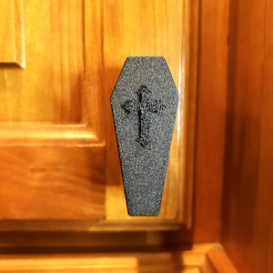 May include: A black, coffin-shaped cabinet pull with a cross design. The pull is mounted on a wooden cabinet with a warm, orange-brown finish. The coffin shape and cross suggest a gothic or Halloween theme.