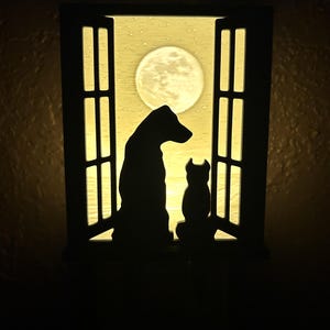 May include: A silhouette of a dog and cat looking out a window at a full moon. The window is made of wood and has a white frame. The moon is a bright yellow color.