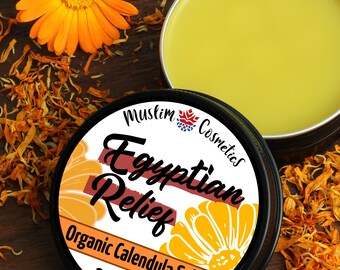 Könnte beinhalten: Eine runde schwarze Dose mit Egyptian Relief Bio-Ringelblumensalbe mit einem weißen Etikett mit dem Text Muslim Cosmetics, Egyptian Relief, Organic Calendula Salve und 2oz / 60ml. Die Dose enthält einen gelben Balsam neben einer orangefarbenen Blume.
