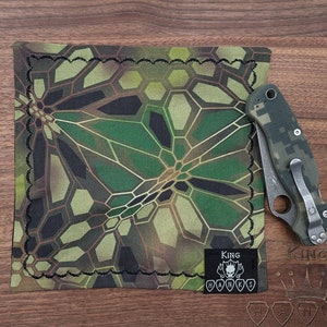 May include: A green and brown camouflage patterned fabric square with a black stitched border. The fabric has a geometric pattern. A black patch with the text "KING HANKS" is sewn on the bottom right corner of the fabric.