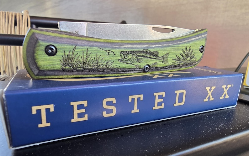 May include: A folding pocket knife with a green and black handle featuring an engraved bass fish design. The blade is silver, and the knife rests on a blue box with the text "TESTED XX" in gold letters. A fishing theme.