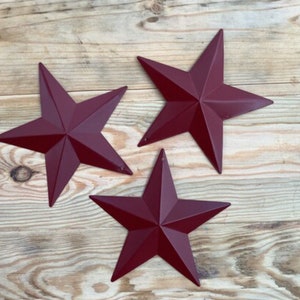 May include: Three burgundy five-pointed stars on a wood background. The stars are made of metal and have a rustic look.