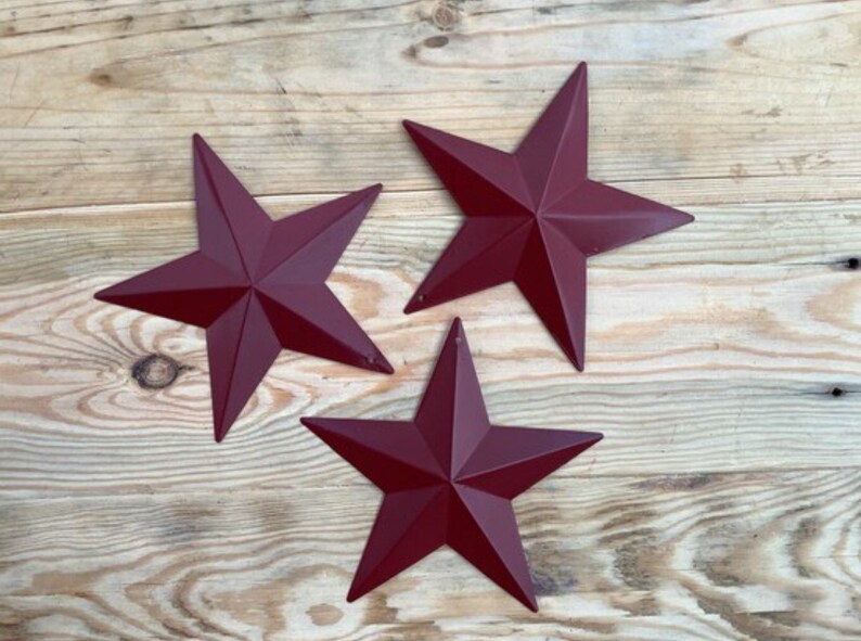 May include: Three burgundy five-pointed stars on a wood background. The stars are made of metal and have a rustic look.