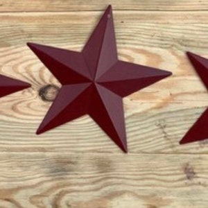 May include: Three burgundy metal star-shaped ornaments. The stars are arranged horizontally on a wood background.
