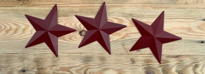May include: Three burgundy metal star-shaped ornaments. The stars are arranged horizontally on a wood background.