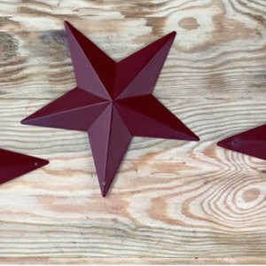 May include: Three burgundy metal star shapes on a wood background. The stars are five-pointed and have a rustic look.