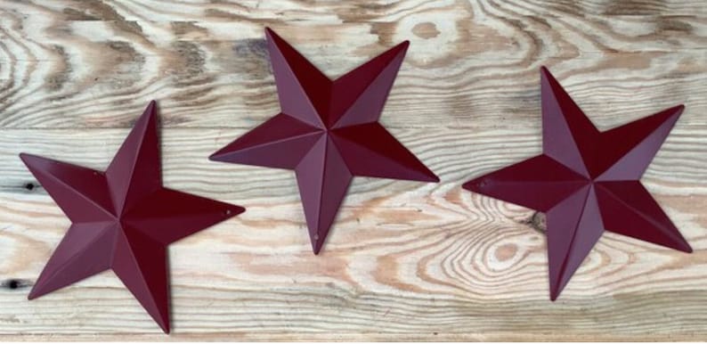 May include: Three burgundy metal star shapes on a wood background. The stars are five-pointed and have a rustic look.