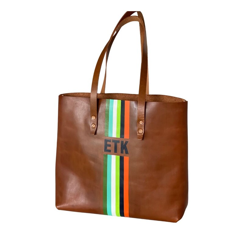 May include: A brown leather tote bag with vertical stripes in green, white, orange, and teal. The bag has brown leather handles and the initials "ETK" in black. A stylish accessory.