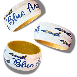May include: Three white wooden bangles with gold interiors. Each bangle features the words "Blue Angels" in blue script and illustrations of blue and yellow jets. The background has a cloudy design.