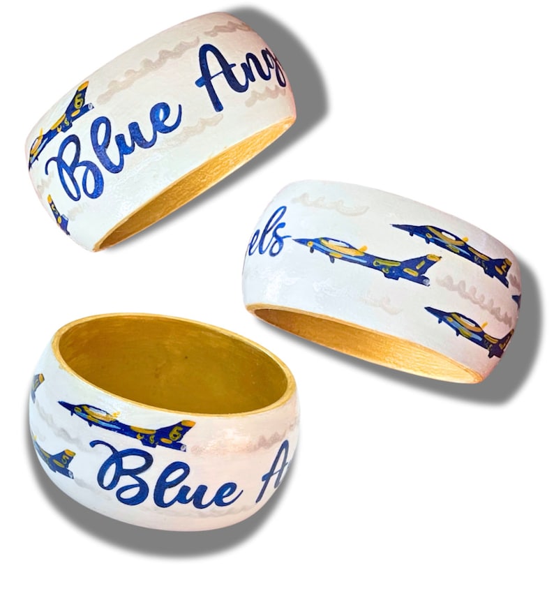 May include: Three white wooden bangles with gold interiors. Each bangle features the words "Blue Angels" in blue script and illustrations of blue and yellow jets. The background has a cloudy design.