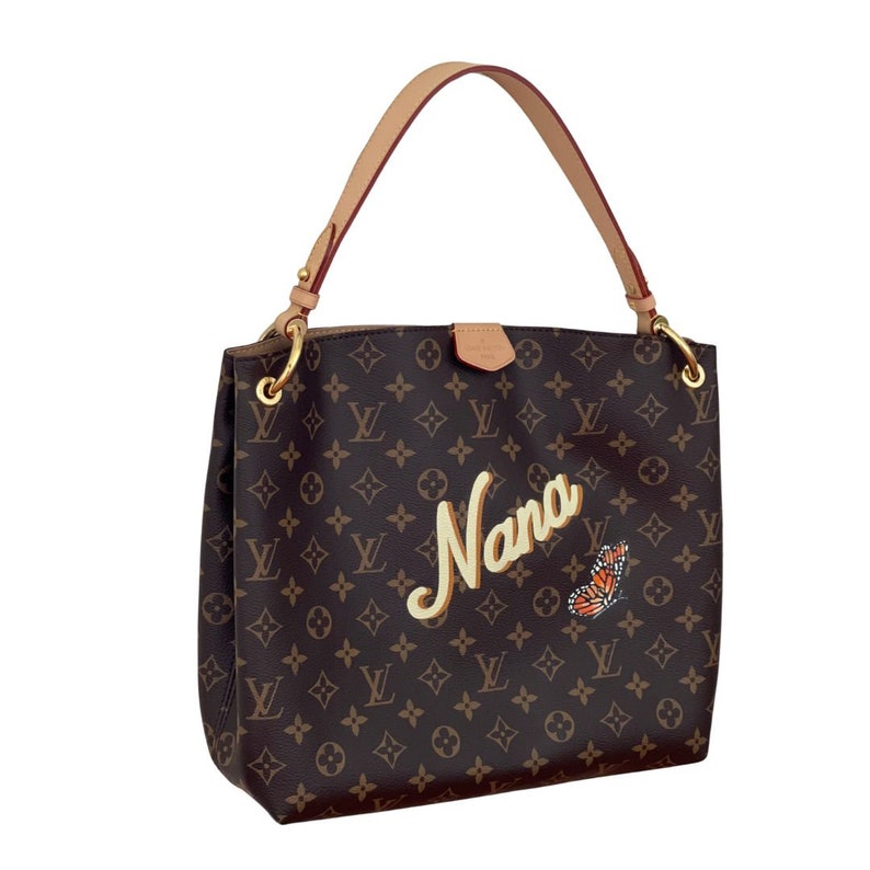 May include: Brown monogrammed handbag with a tan leather handle and gold-tone hardware. The word "Nana" is written in a cursive font, with a butterfly graphic. The bag has a shoulder strap.