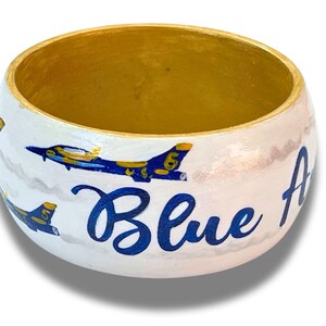 May include: A white wooden bowl with a gold interior and the words "Blue Angels" in blue script. The bowl is decorated with blue and yellow airplanes and gray clouds.