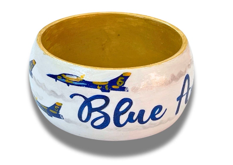 May include: A white wooden bowl with a gold interior and the words "Blue Angels" in blue script. The bowl is decorated with blue and yellow airplanes and gray clouds.