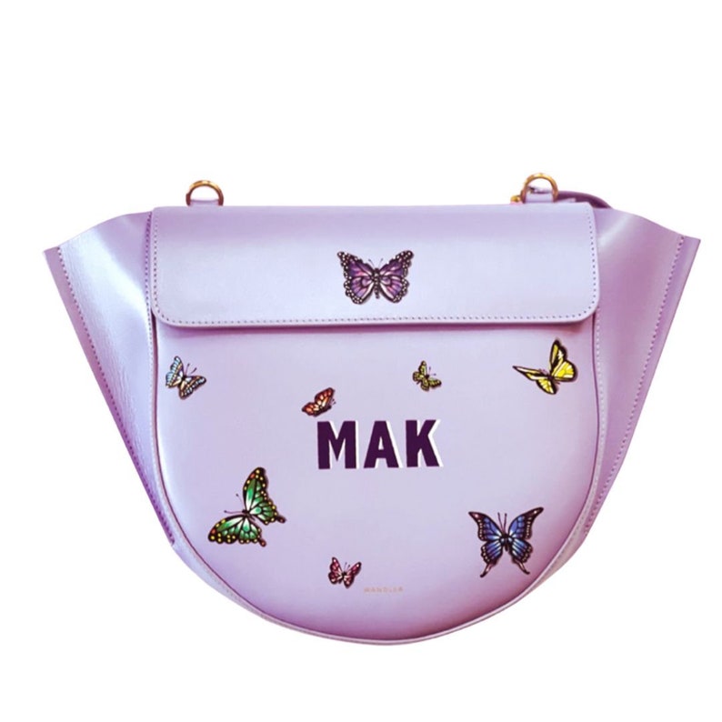 May include: A lilac handbag with a flap closure and a curved base. The bag is adorned with colourful butterfly appliqu&eacute;s and the letters "MAK" in silver. It features a gold-coloured shoulder strap.