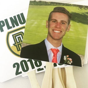 May include: A white and green fan with the text "PLNU 2018" and a photo of a man in a black suit and a red tie. The fan is made of paper and has a wooden handle.