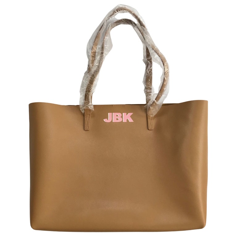 May include: A tan tote bag with a rectangular shape and two handles. The bag features the initials "JBK" in pink letters on the front. The handles are wrapped in clear plastic.