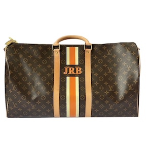 May include: Brown monogrammed duffel bag with a vertical orange and white stripe down the centre. The bag has tan leather accents, a top handle, and the initials "JRB" in the centre of the stripe.