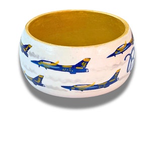 May include: A hand-painted wooden bangle bracelet with a white exterior and gold interior. The bracelet features blue and yellow jets with the text "Blue" in cursive. The jets are painted on a white background with gray clouds.